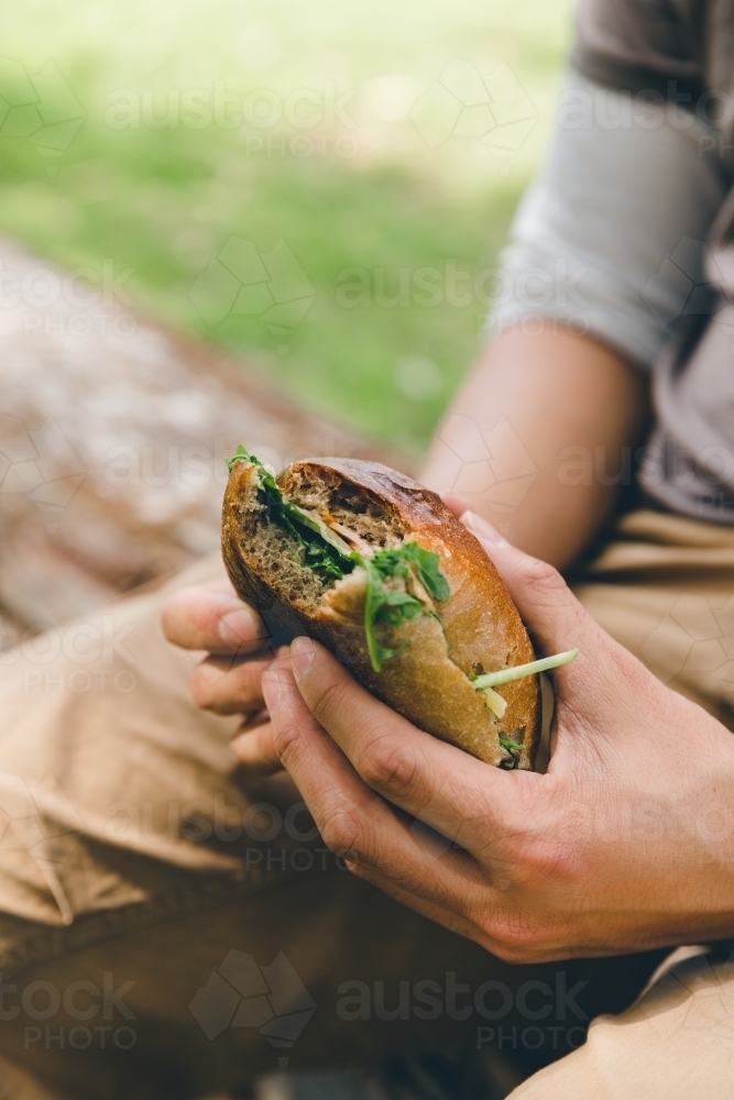 Man eating bread roll sandwich, sitting on log in green park - Australian Stock Image