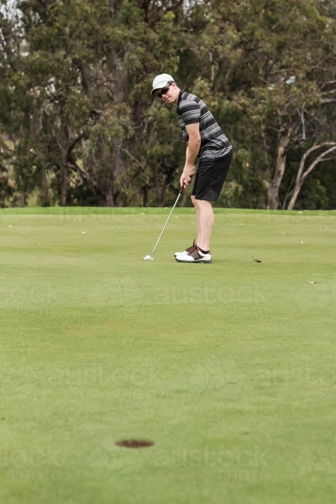 Man holding golf club putter hitting ball into hole on green - Australian Stock Image