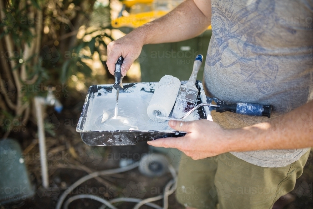 Man holding paint tray with roller and pain brush mixing paint - Australian Stock Image