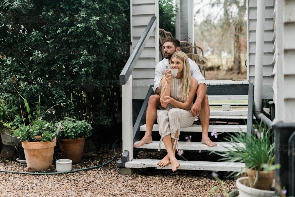 Man holding woman from behind as they sit on the steps of their porch - Australian Stock Image