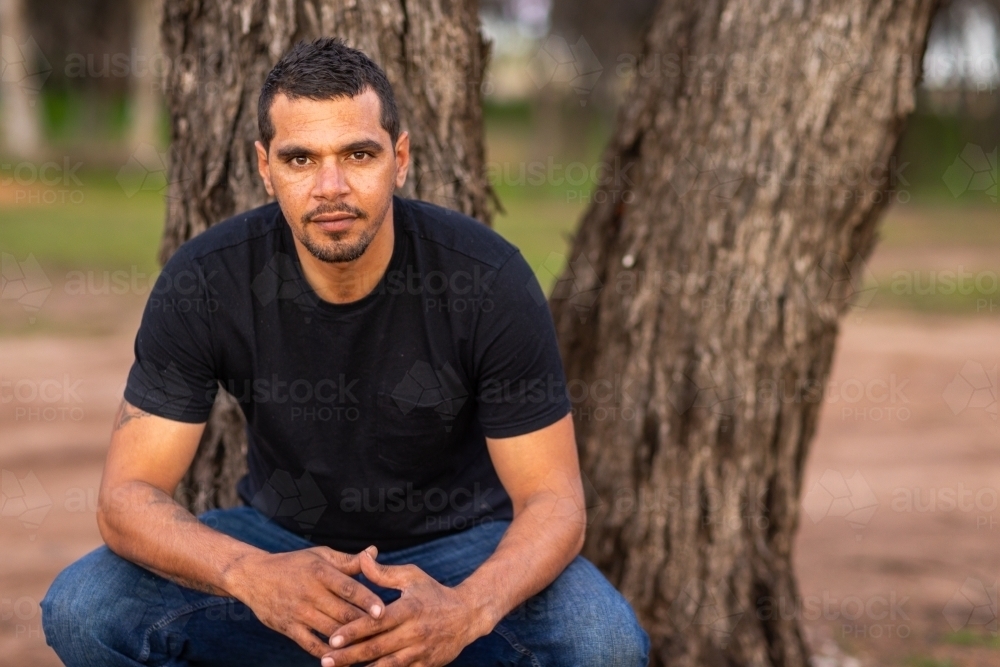 man in black tee shirt squatting beside tree - Australian Stock Image