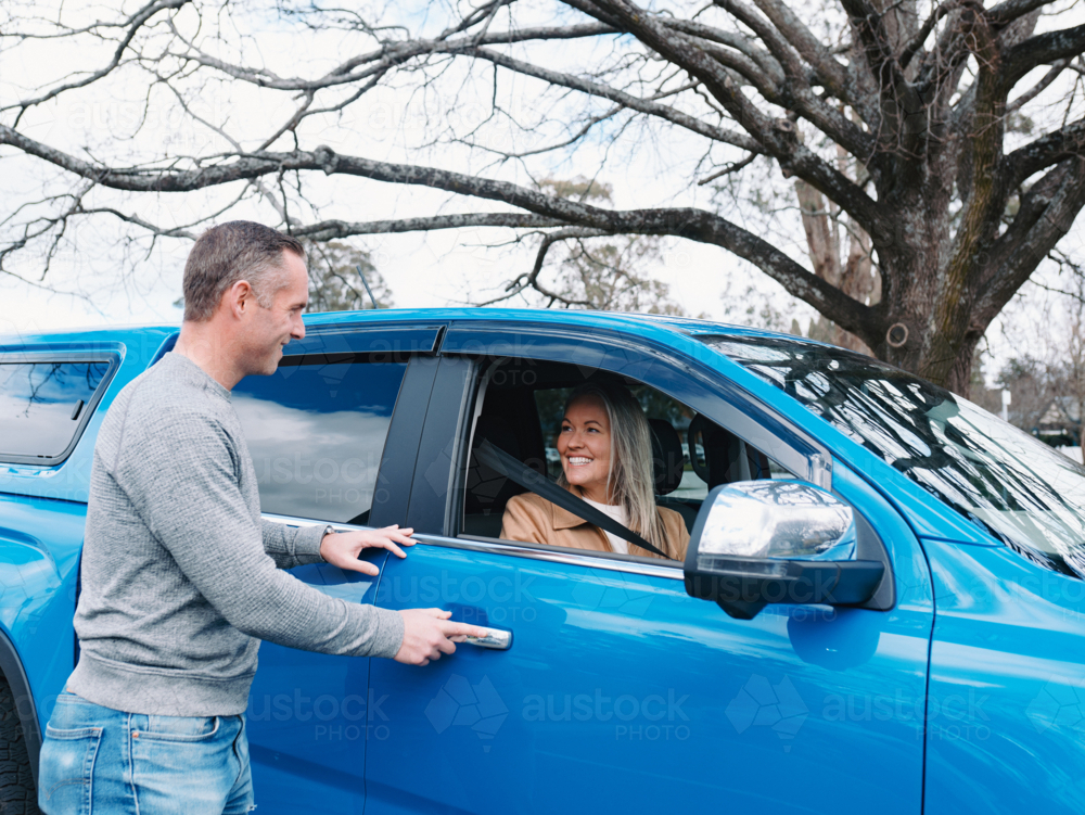 Man in grey sweater closing car door with woman sitting on passenger seat - Australian Stock Image