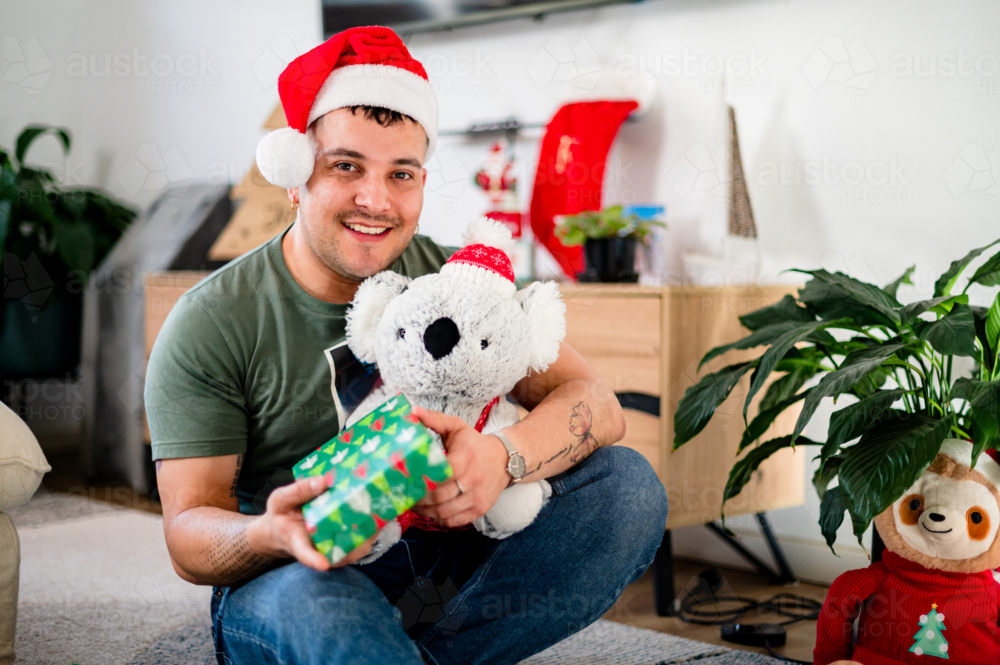 Man in his 30's with colourful gift wearing a Santa hat, smiling amidst decorations - Australian Stock Image