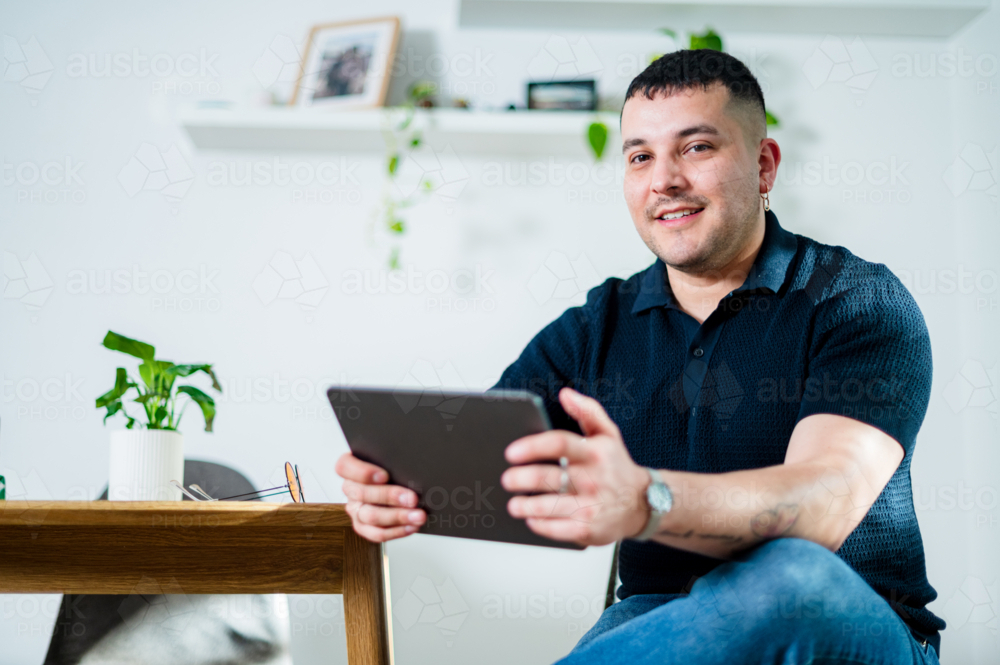 Man in his 30s sits comfortably at home, engaging with a tablet in a relaxed setting. - Australian Stock Image