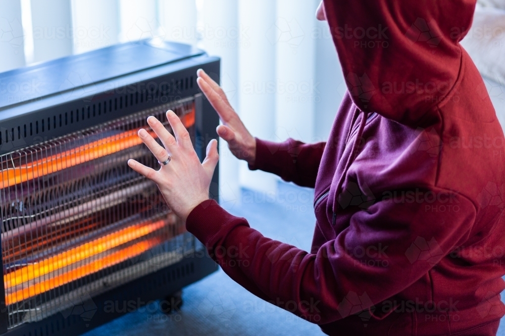 Man in hoodie jumper warming up his cold hand in front of space heater inside home - Australian Stock Image