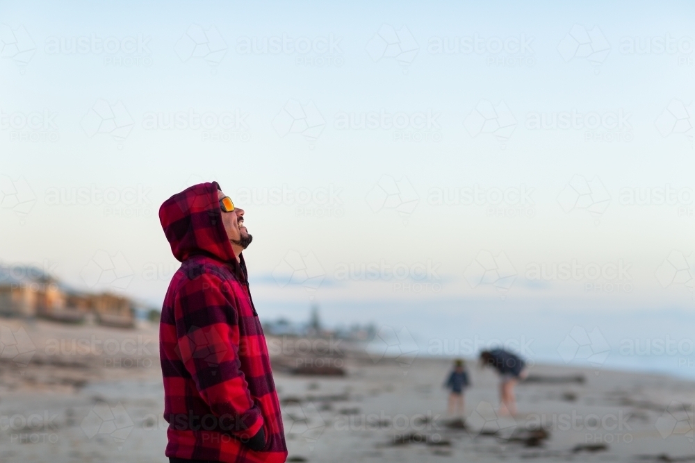 man in red hoody on beach with mother and child blurred in background - Australian Stock Image