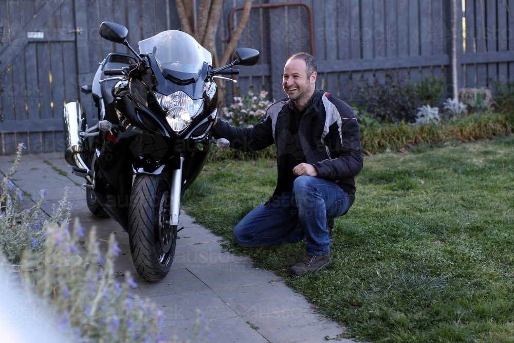 Man laughing and crouching next to motorbike in garden - Australian Stock Image