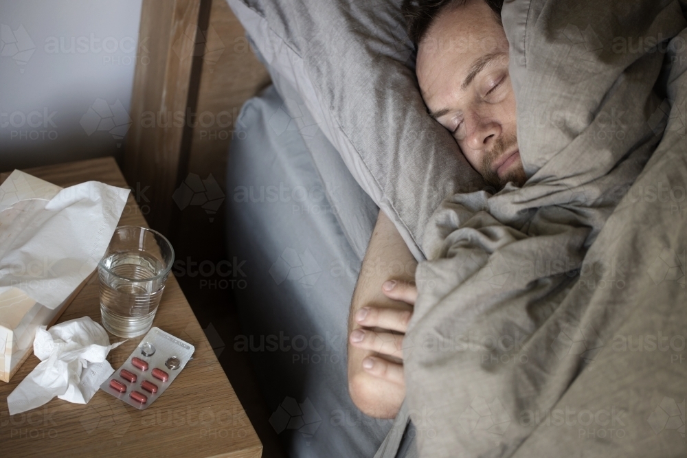 Man lying sick in bed with tissue box, medicine and glass of water - Australian Stock Image