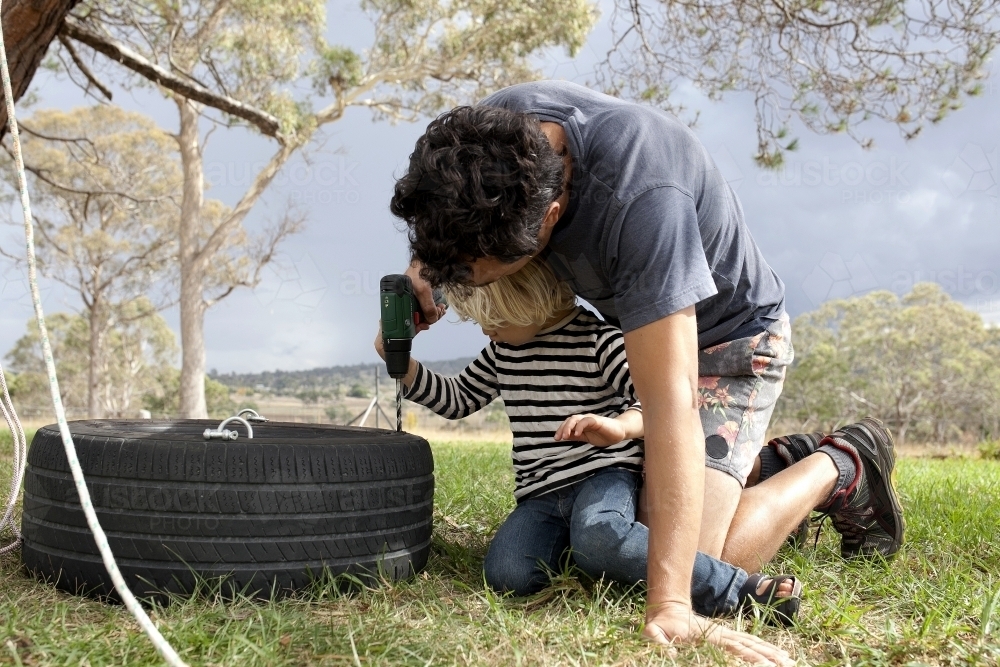 Man making a tyre swing in country backyard with help from young boy - Australian Stock Image