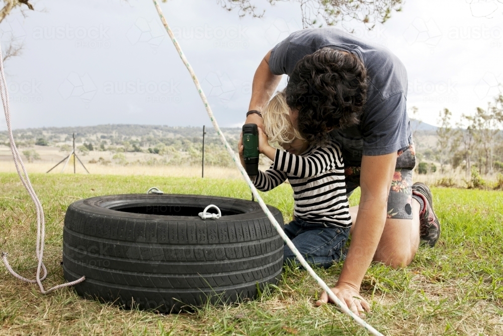 Man making a tyre swing with help from young boy in country back yard - Australian Stock Image