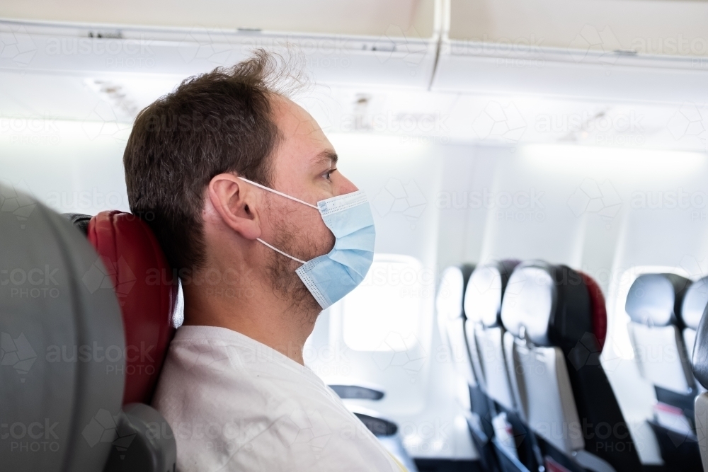 Man on an airplane wearing a surgical mask traveling during the COVID-19 coronavirus pandemic - Australian Stock Image
