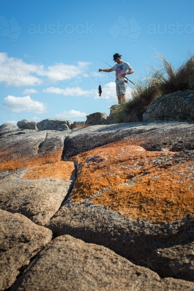 Man on boulder rocks at beach holding a fresh caught fish - Australian Stock Image
