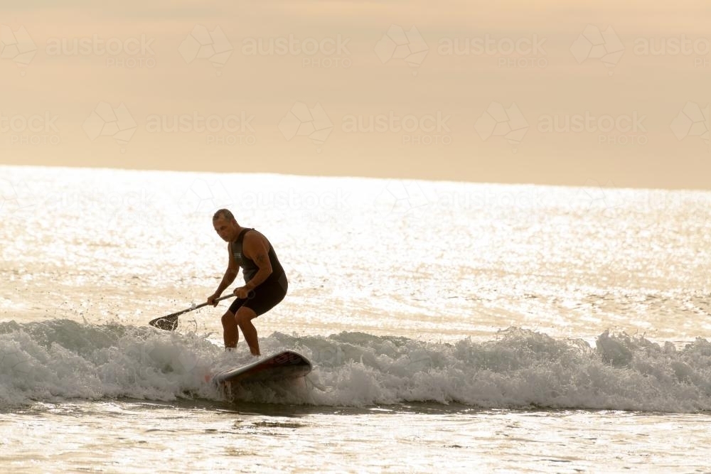 Man on paddleboard riding wave and backlit by dawn light - Australian Stock Image