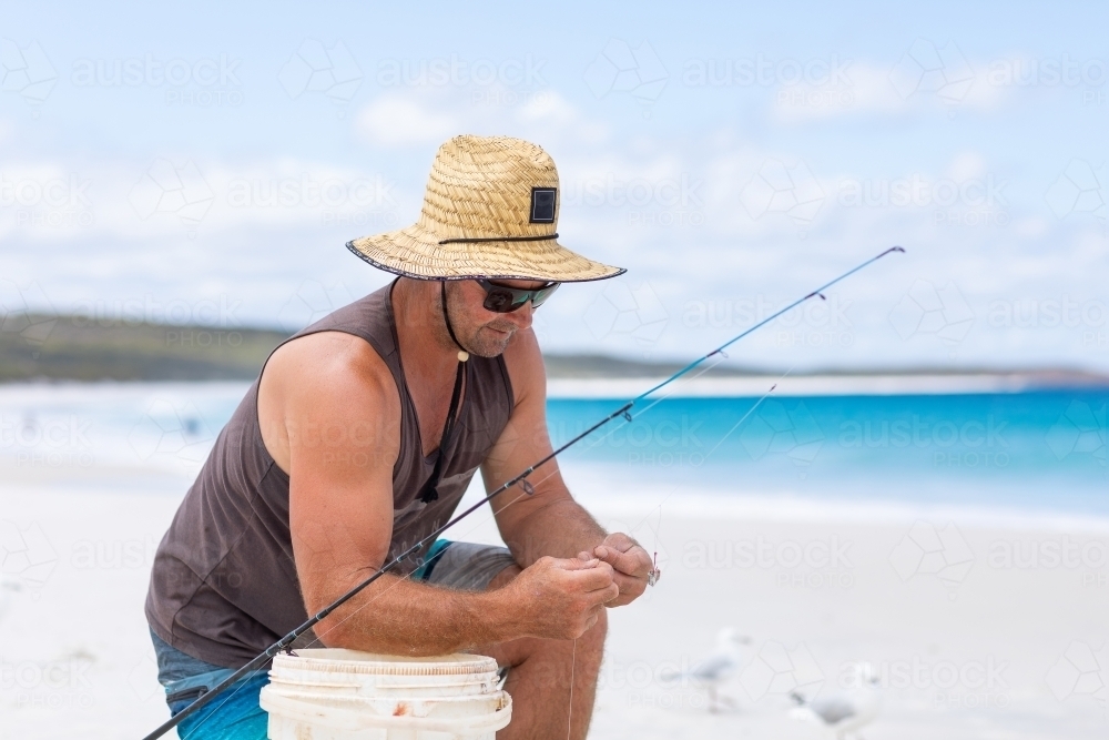 man on white sand beach kneeling down baiting fishing line - Australian Stock Image