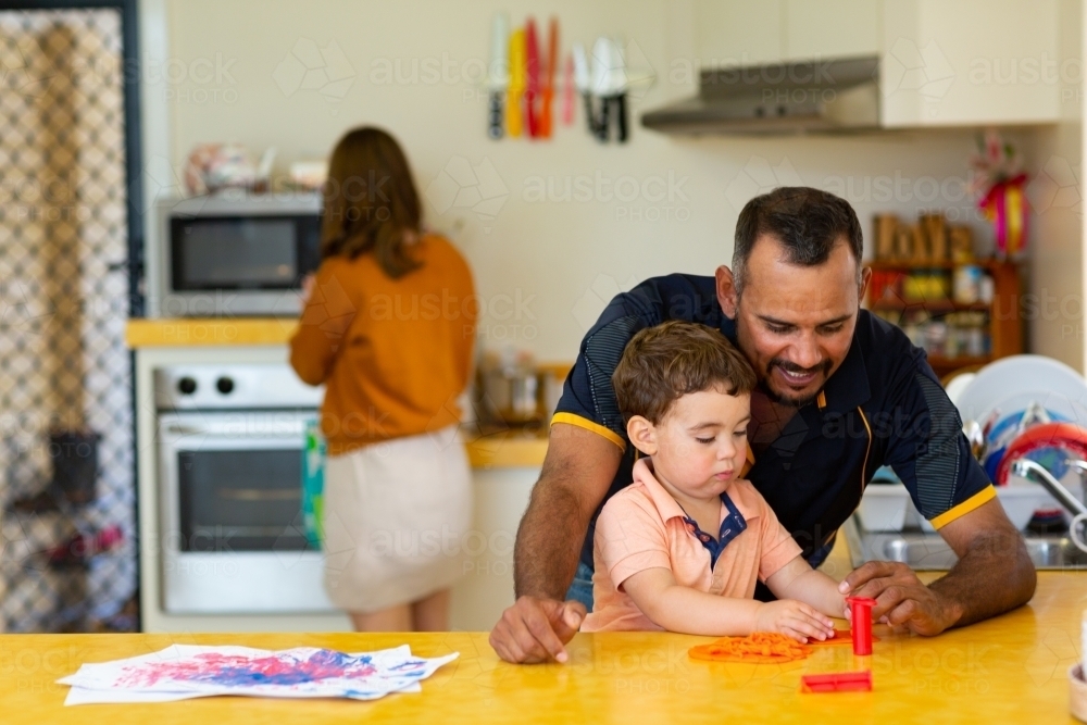 man playing with toddler in kitchen with woman in background - Australian Stock Image
