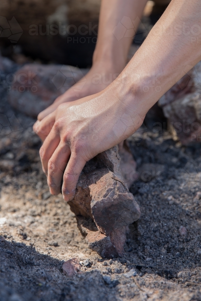 Man preparing camp fire by clearing away ash and charcoal with rock - Australian Stock Image
