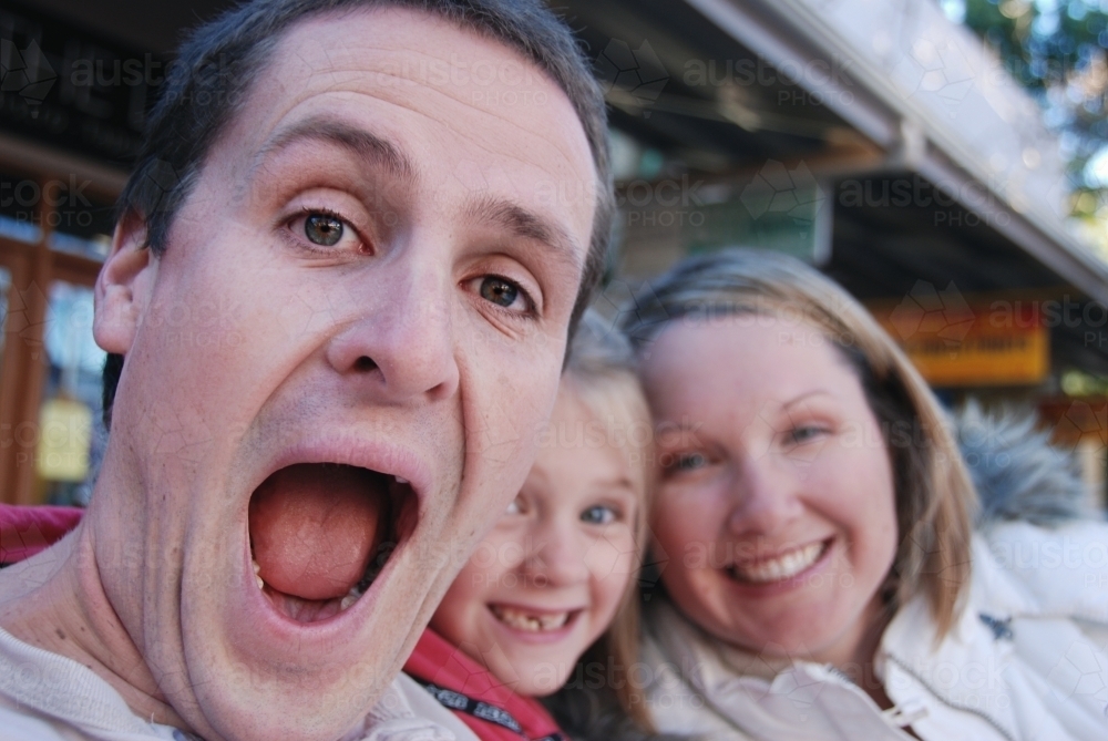 Man pulling a crazy face to camera, with wife and child in the background - Australian Stock Image