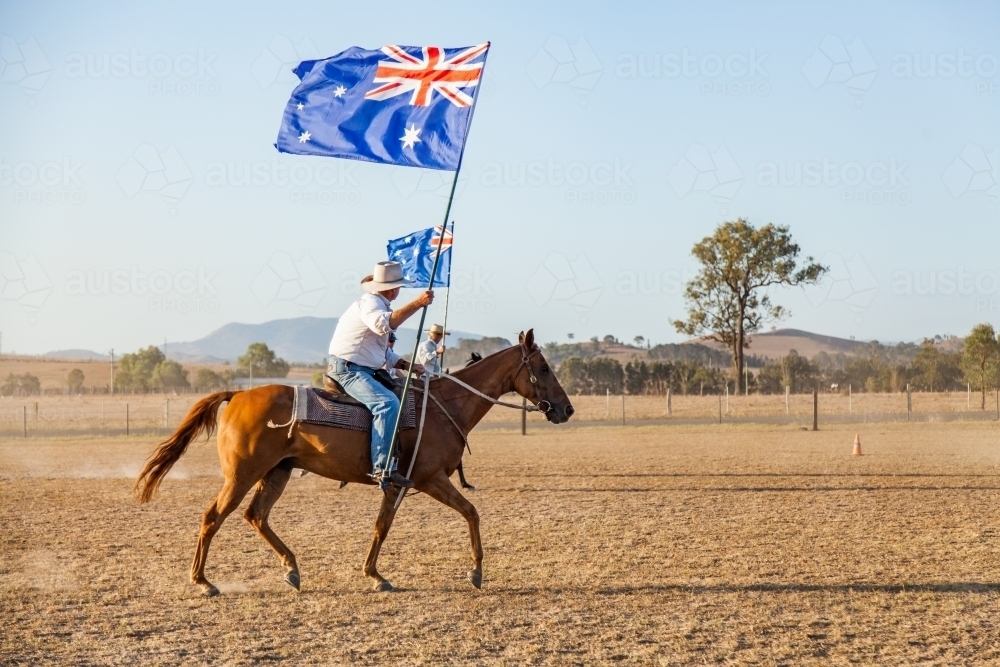 Man riding Australian stock horse with flag in dusty paddock - Australian Stock Image
