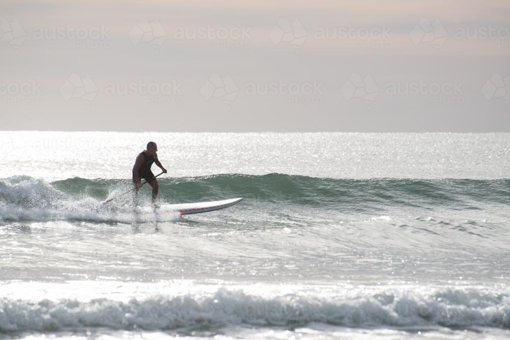 Man riding wave on paddleboard in glistening ocean - Australian Stock Image