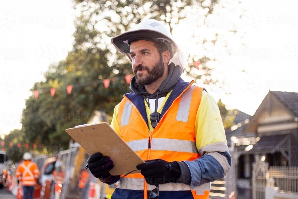 man road worker with beard wearing white hat and orange and yellow jacket holding his notes - Australian Stock Image