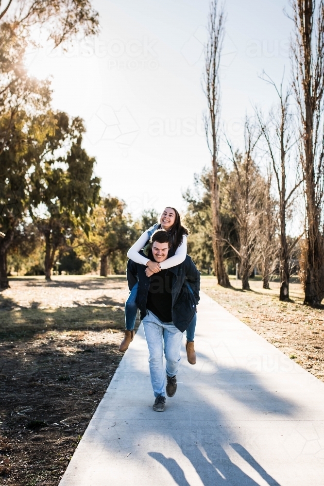 Man running carrying girlfriend piggy back laughing on path in park - Australian Stock Image
