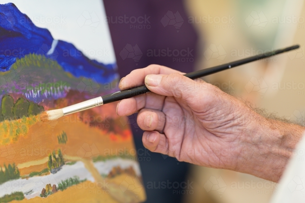 Man's hand holding paintbrush with painting in background - Australian Stock Image