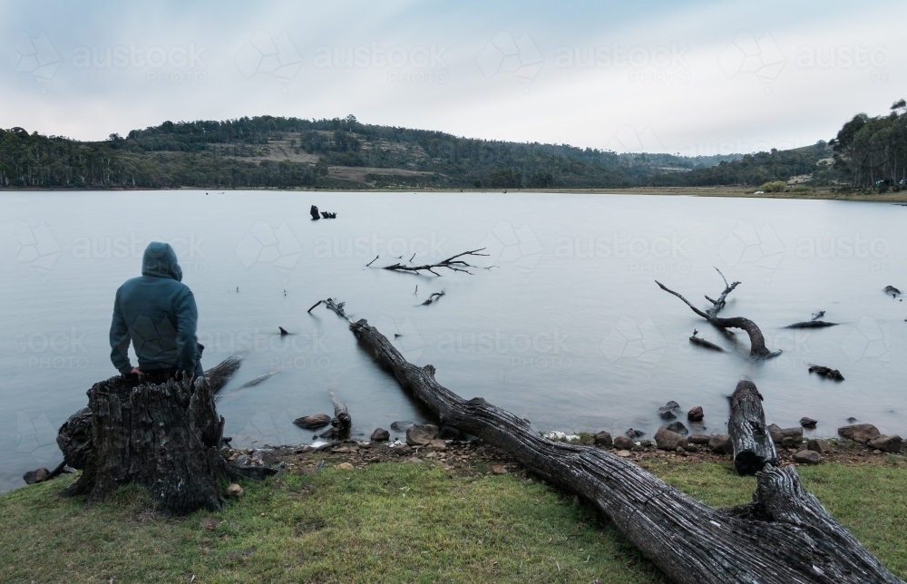 Man Seated on a Tree Stump on Lake Shore, Tasmania - Australian Stock Image