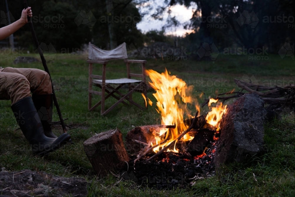 Man sitting beside roaring campfire at dusk - Australian Stock Image