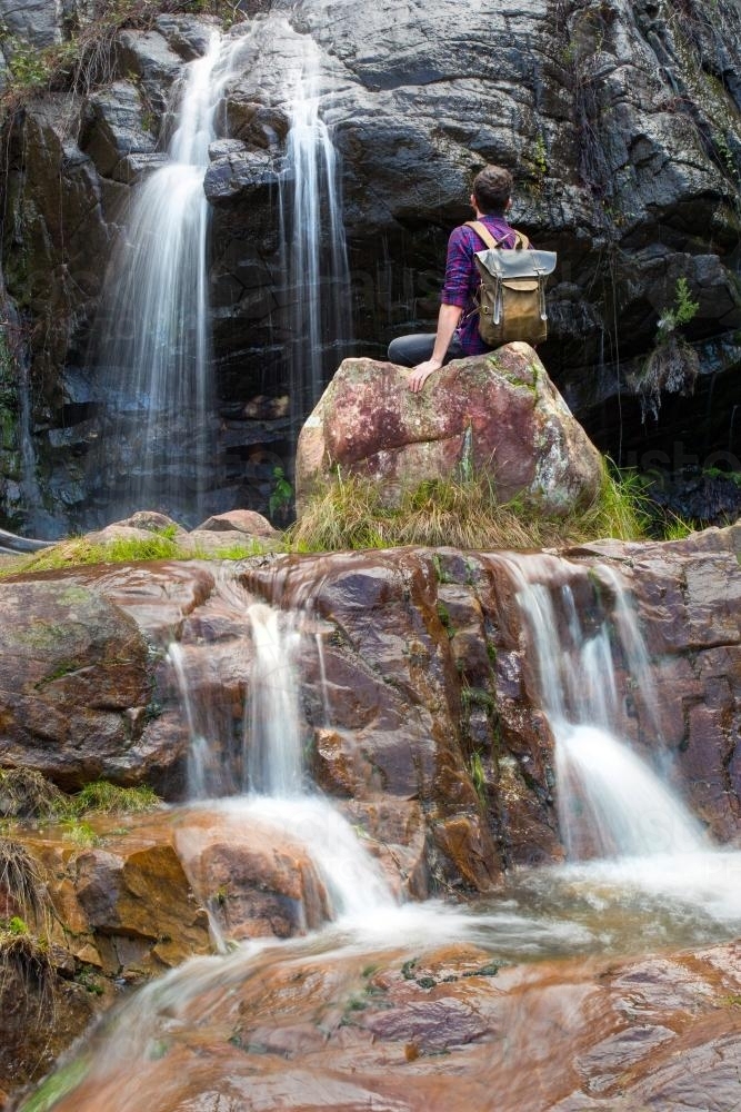 Man sitting by cascading waterfall - Australian Stock Image