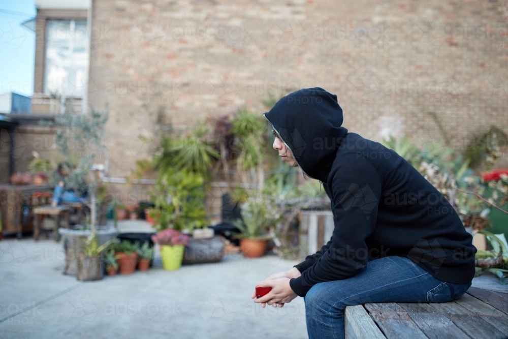 Man sitting in courtyard wearing black hoodie and sunglasses - Australian Stock Image