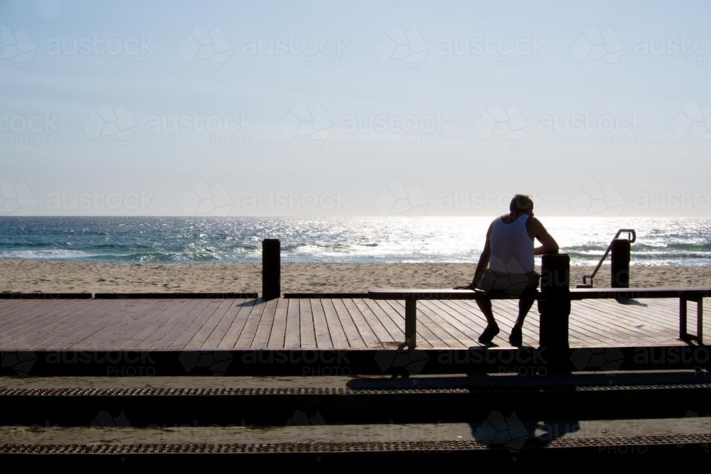 Man sitting on a bench silhouetted against the sun and sea - Australian Stock Image