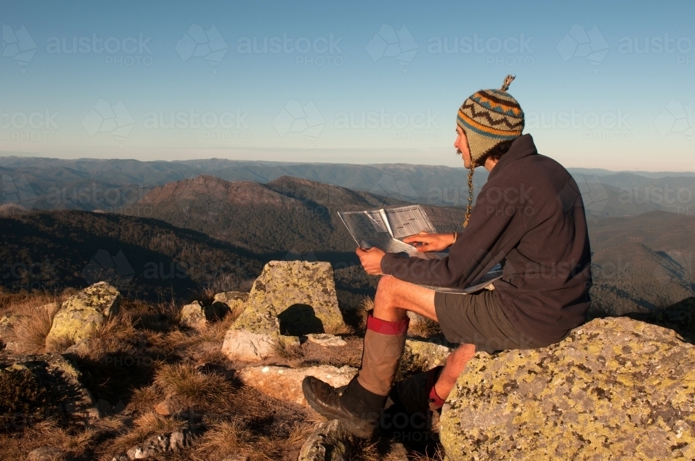 Man sitting on a rock overlooking mountain ranges, looking at a map - Australian Stock Image
