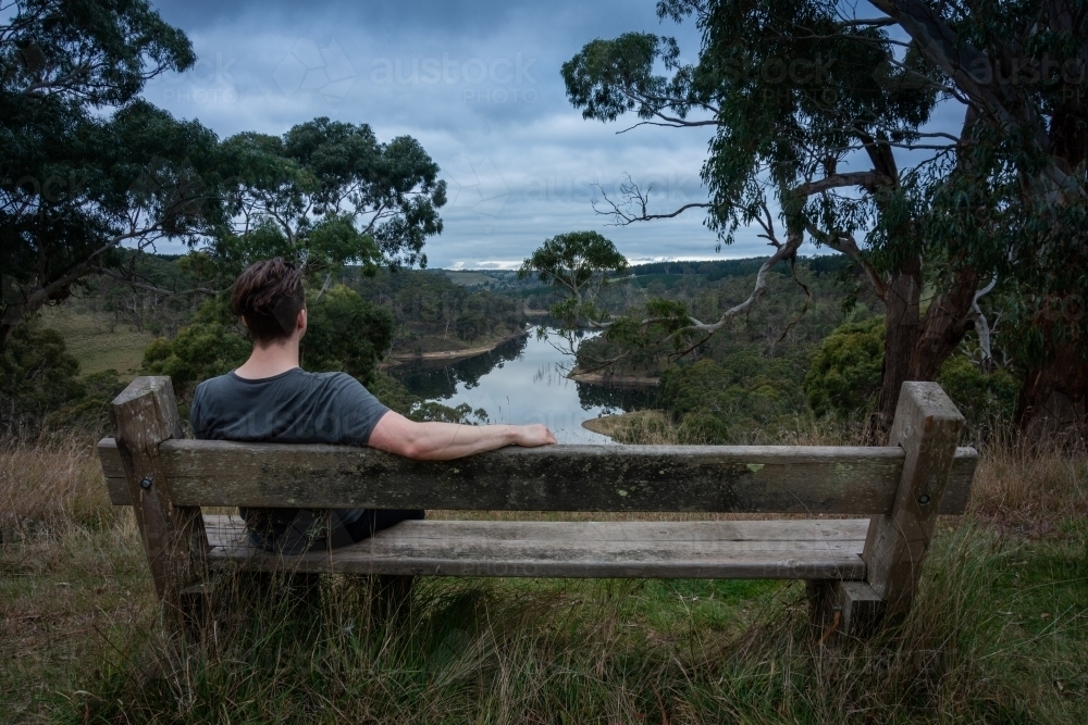 Man Sitting on Bench above a lake on a stormy afternoon - Australian Stock Image