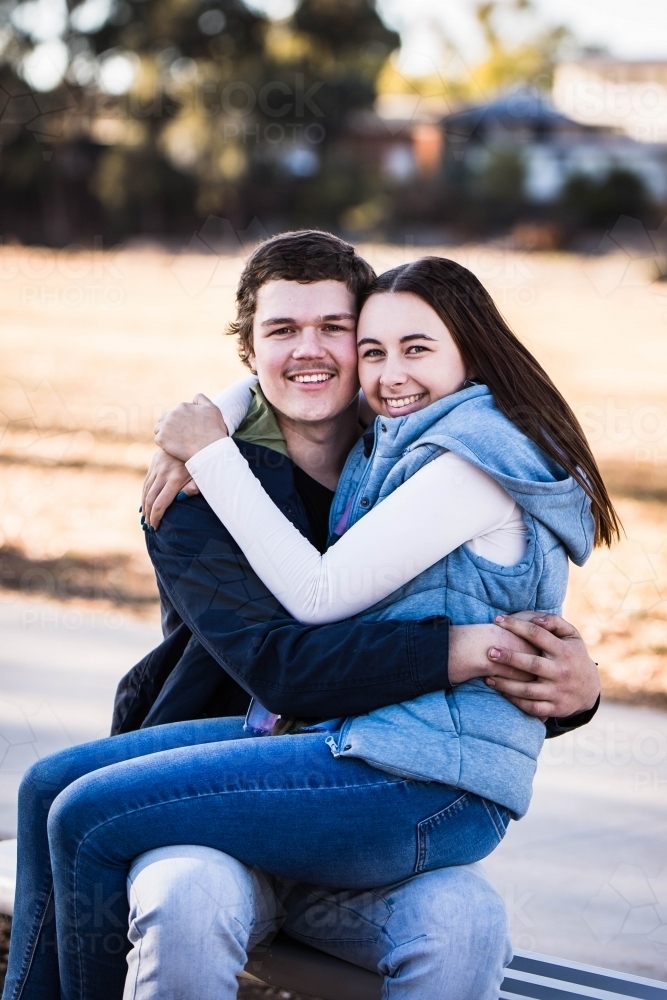 Man sitting on bench with girlfriend on lap with arms around each other smiling - Australian Stock Image