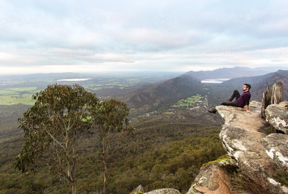 Man sitting on rocks looking out over landscape of green valleys - Australian Stock Image