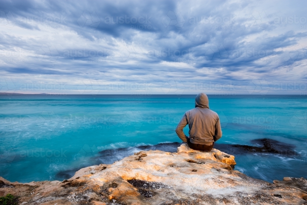Man sitting on the edge of a rock, looking out at turquoise ocean and cloudy sky - Australian Stock Image