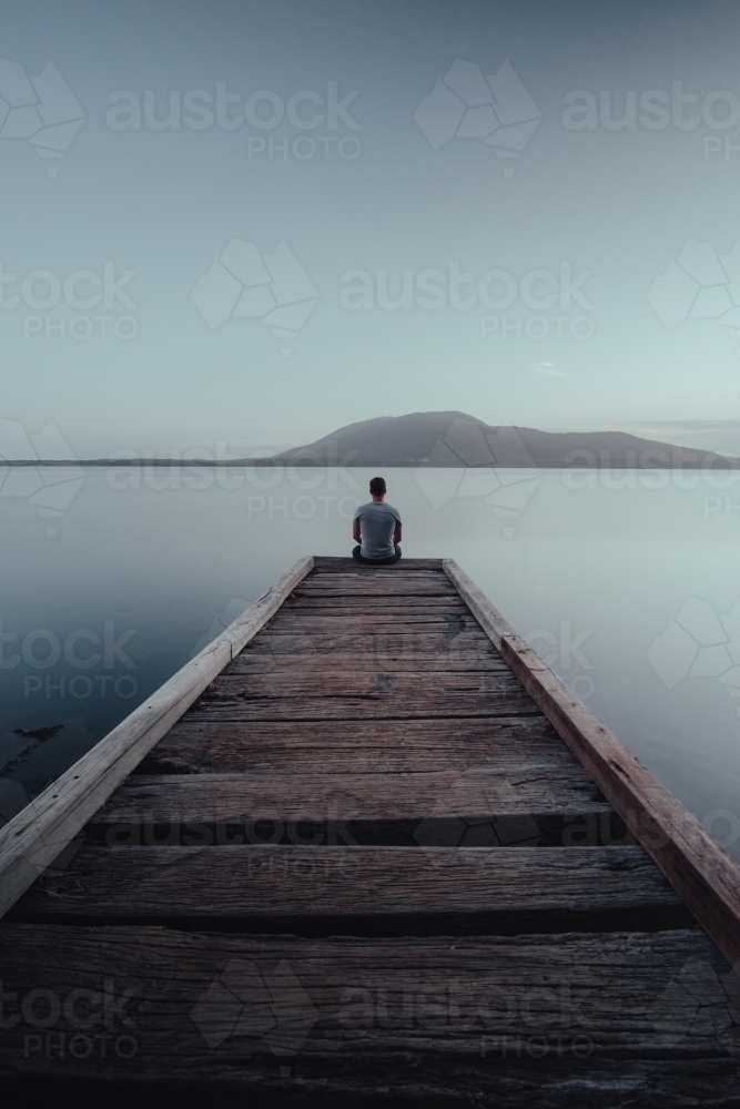 Man sitting on the end of a jetty looking over a lake with a mountain in the background. - Australian Stock Image