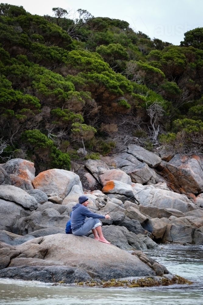 Man sitting with small child behind  and fishing on granite rocks in coastal setting - Australian Stock Image