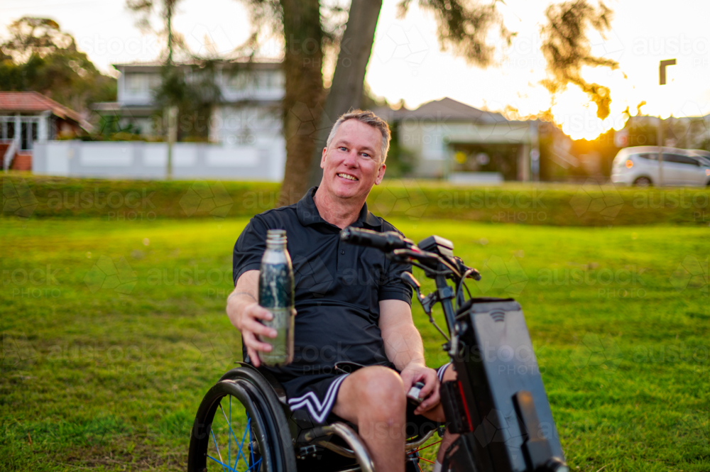 Man smiles while seated in a wheelchair, enjoying a sunny day in a park with greenery. - Australian Stock Image