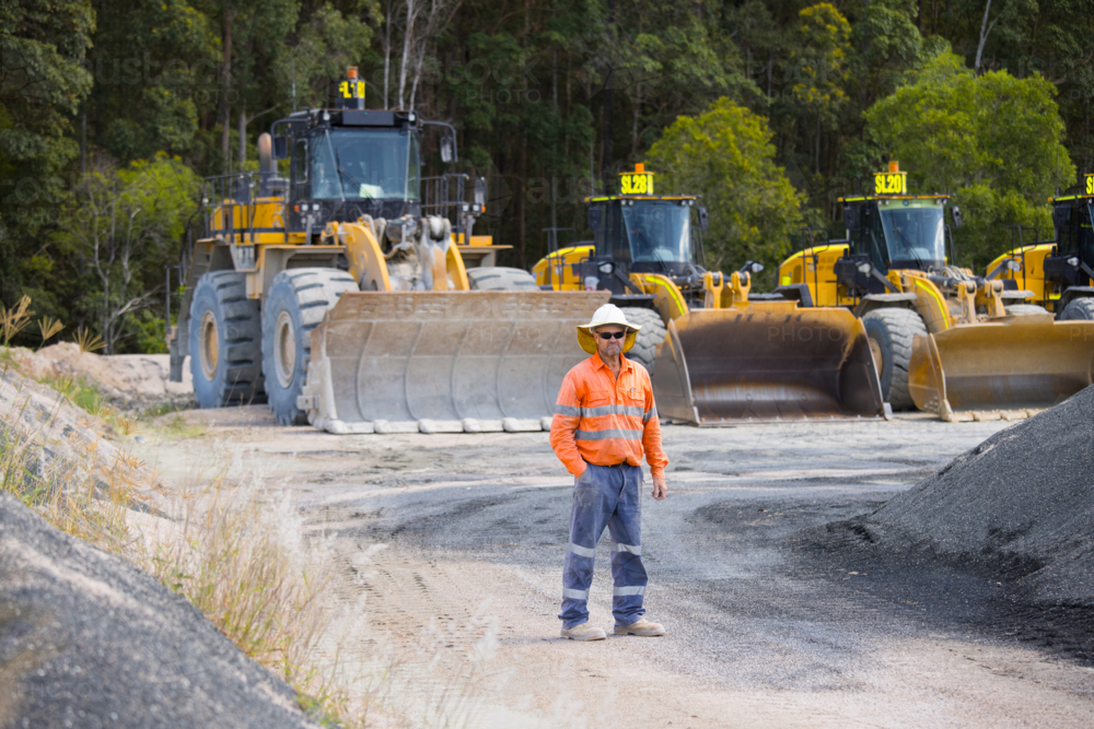 Man standing in the middle of the dirt path with payloaders at the back. - Australian Stock Image