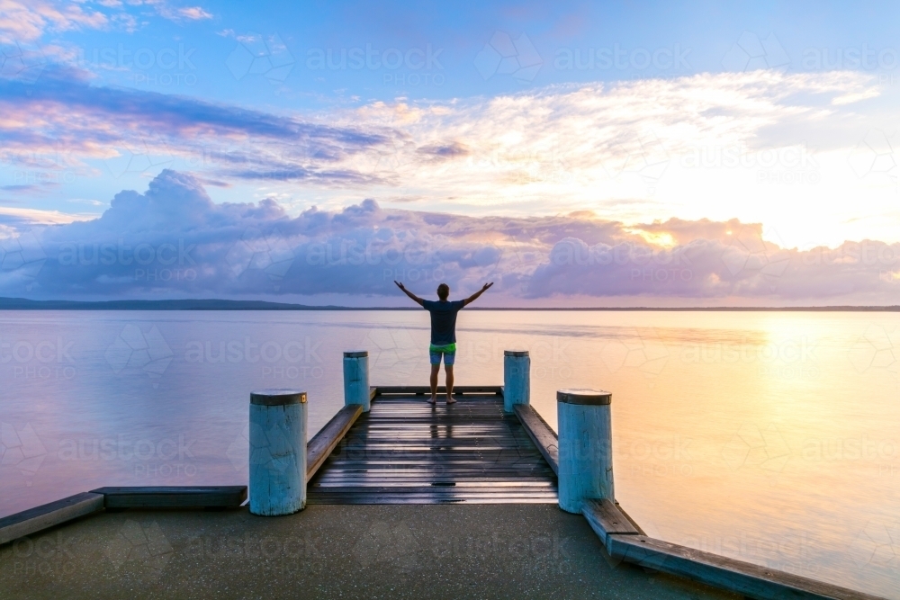 Man standing with outstretched arms on a jetty at sunrise - Australian Stock Image