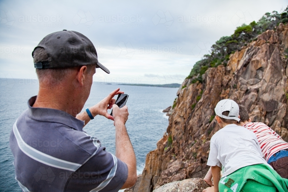 Man taking photos of his boys looking down cliff to the sea - Australian Stock Image