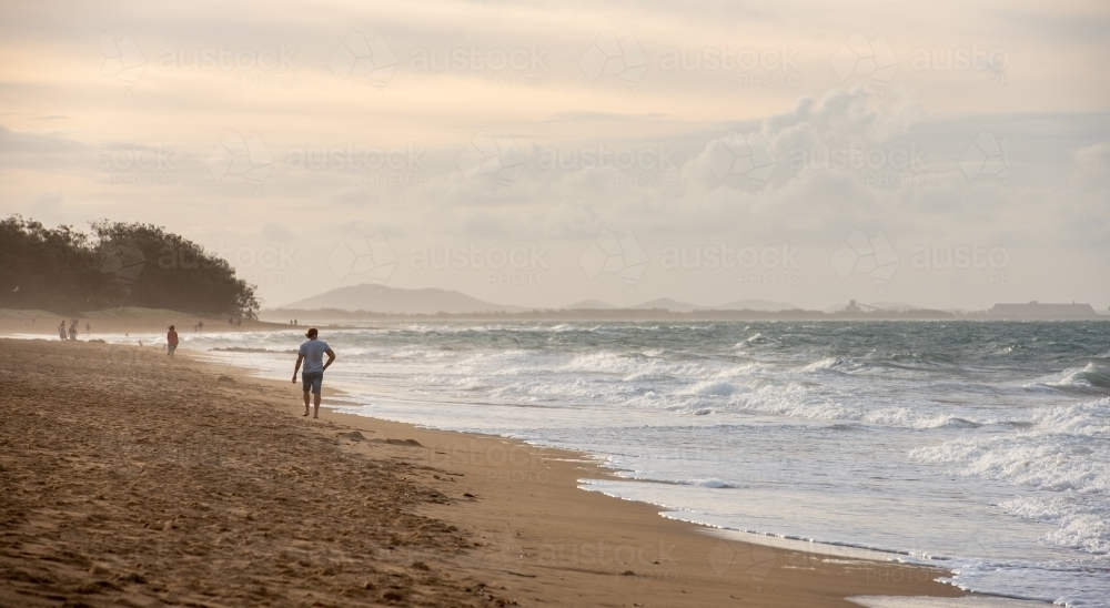 Man walking along Tannum Sands beach with industry in the background, Gladstone Region, Queensland - Australian Stock Image
