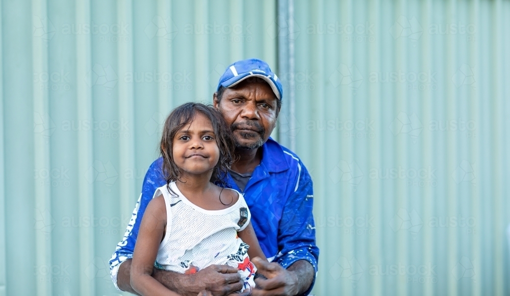 man with arms around little kid sitting on his knee in front of tin fence - Australian Stock Image