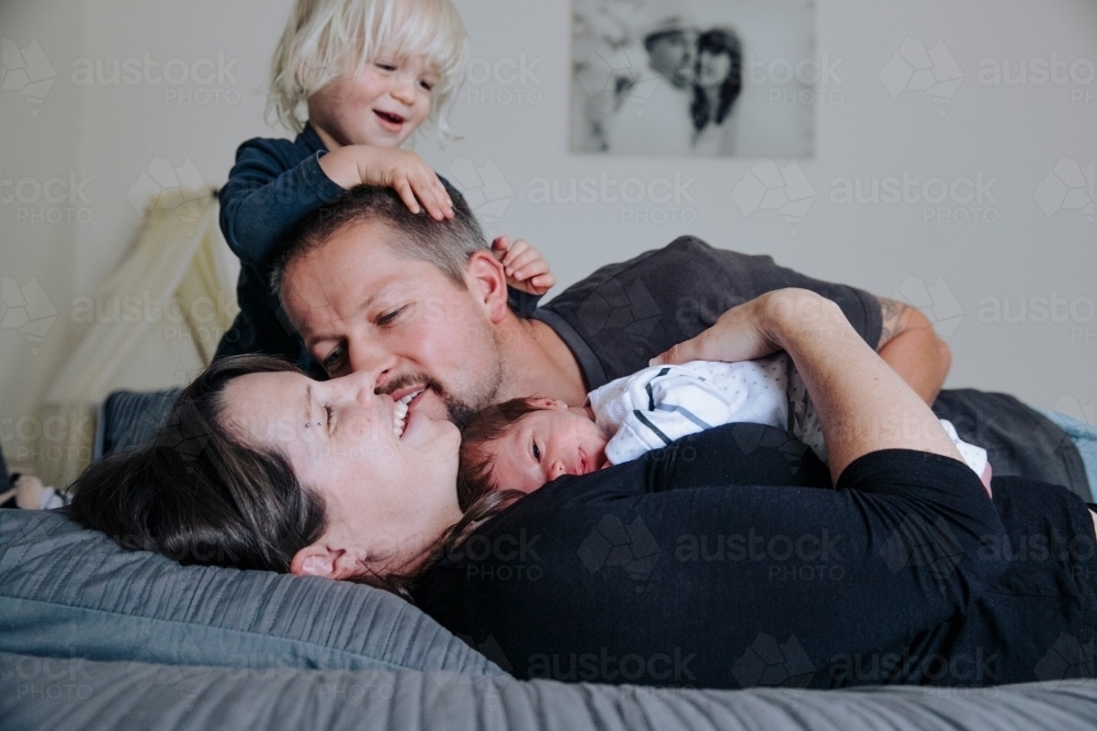 Married couple with their toddler and newborn, lying on the bed with wedding photo on the wall - Australian Stock Image
