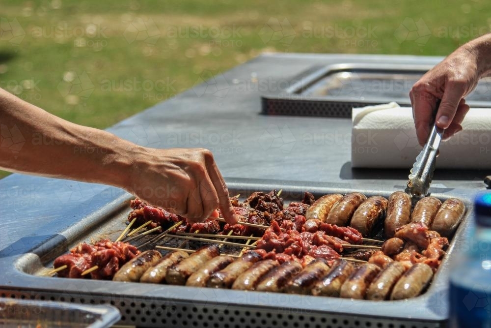 Mates cooking sausages and kebabs on a BBQ at the park - Australian Stock Image