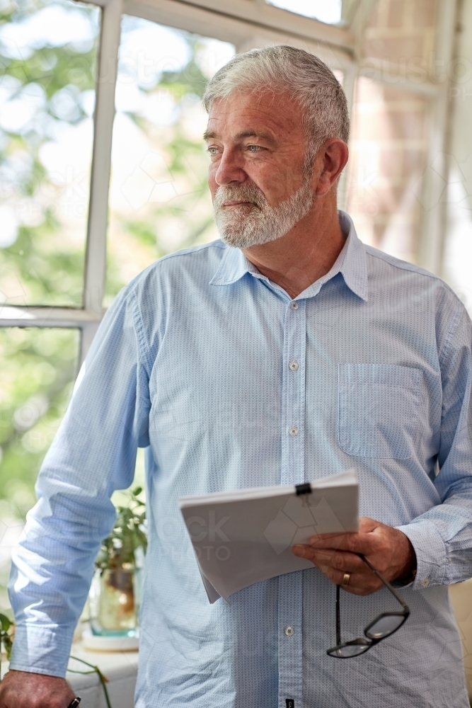 Mature business man holding files while standing, looking our a studio window - Australian Stock Image