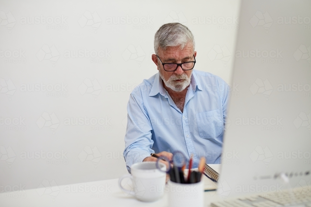 Mature business man sitting at a white desk, working on a computer - Australian Stock Image