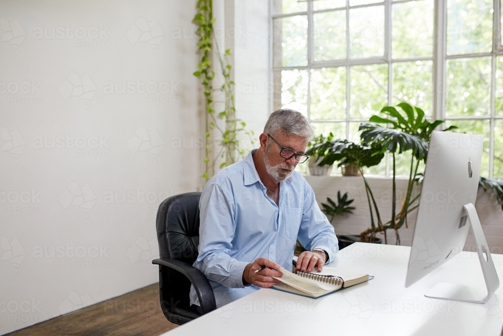 Mature businessman sitting at a desk, reading in an open-plan office - Australian Stock Image