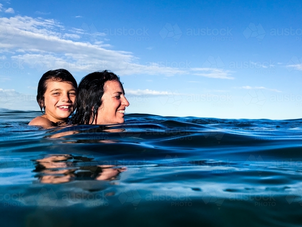 Mature woman and young boy on her back swimming through surface of calm blue ocean - Australian Stock Image